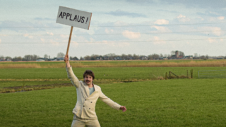 Man in licht pak staat juichend in een uitgestrekt weiland en houdt een bord omhoog met de tekst “APPLAUS!” tegen een achtergrond van Hollandse wolkenlucht en hoogspanningsmast.