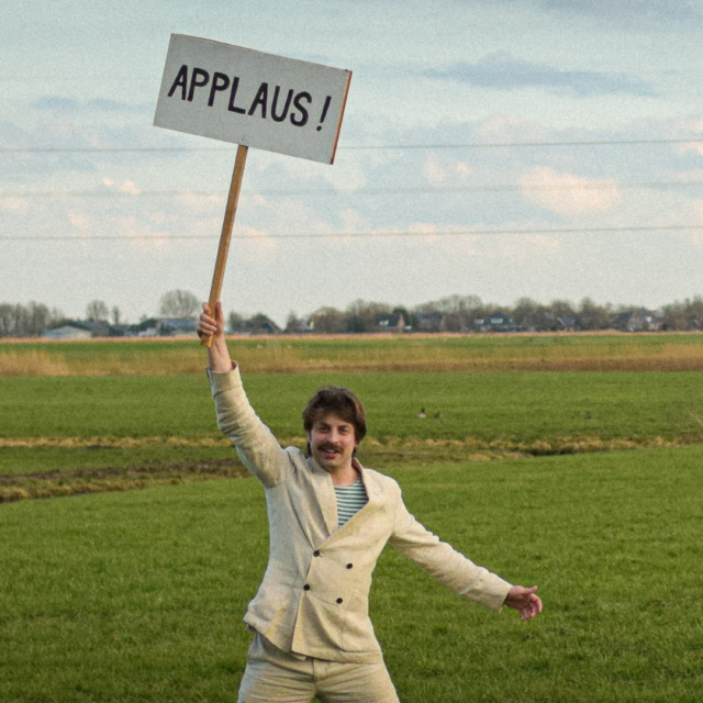 Man in licht pak staat juichend in een uitgestrekt weiland en houdt een bord omhoog met de tekst “APPLAUS!” tegen een achtergrond van Hollandse wolkenlucht en hoogspanningsmast.