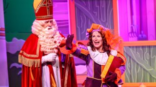Sinterklaas in zijn traditionele rode mantel staat naast een Pietenfiguur, die vrolijk glimlacht en een boek vasthoudt in een kleurrijk decor.