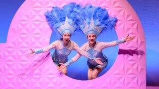 Plien en Bianca staan vrolijk door een roze gat, met een blauw glitterpak aan en blauwe glitterveren op hun hoofd