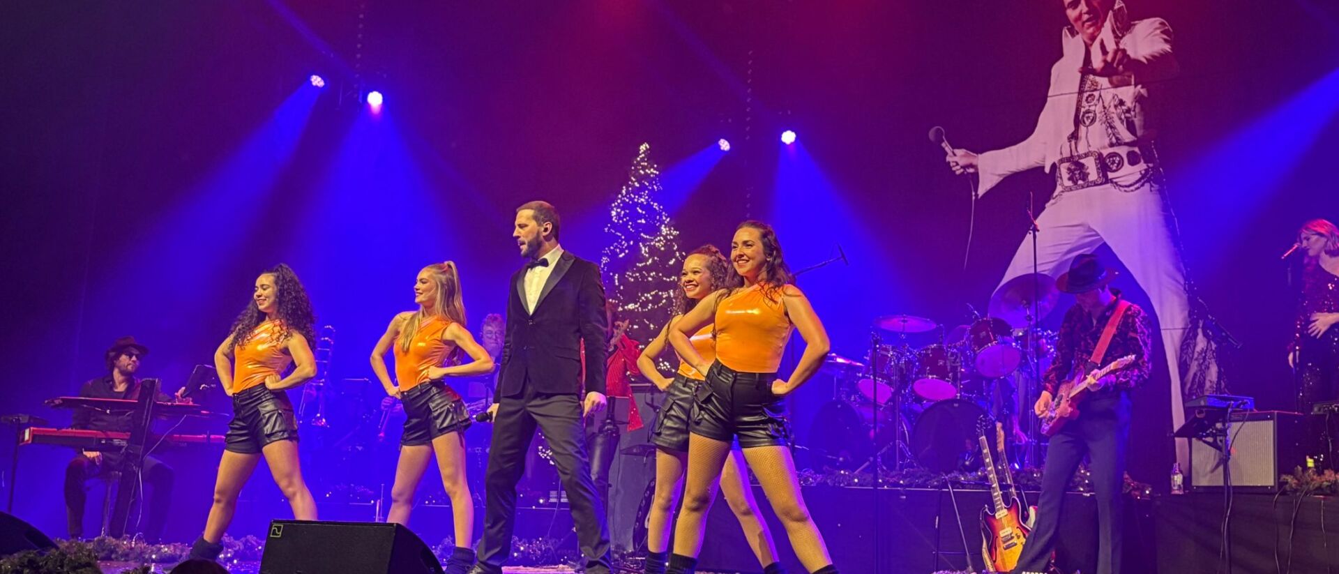 Bouke Scholten tijdens de kerstspecial van Bouke Rocks Elvis met dansers en de ElvisMatters Band, terwijl een groot beeld van Elvis in de achtergrond te zien is
