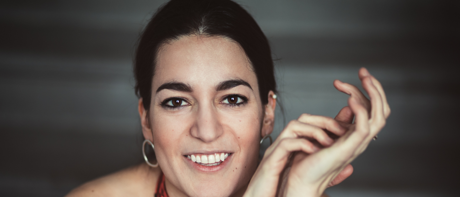 Vrouw met donkere haren en een rode fluwelen top kijkt lachend in de camera, haar handen speels gevouwen voor zich, met een zachte, donkere achtergrond.