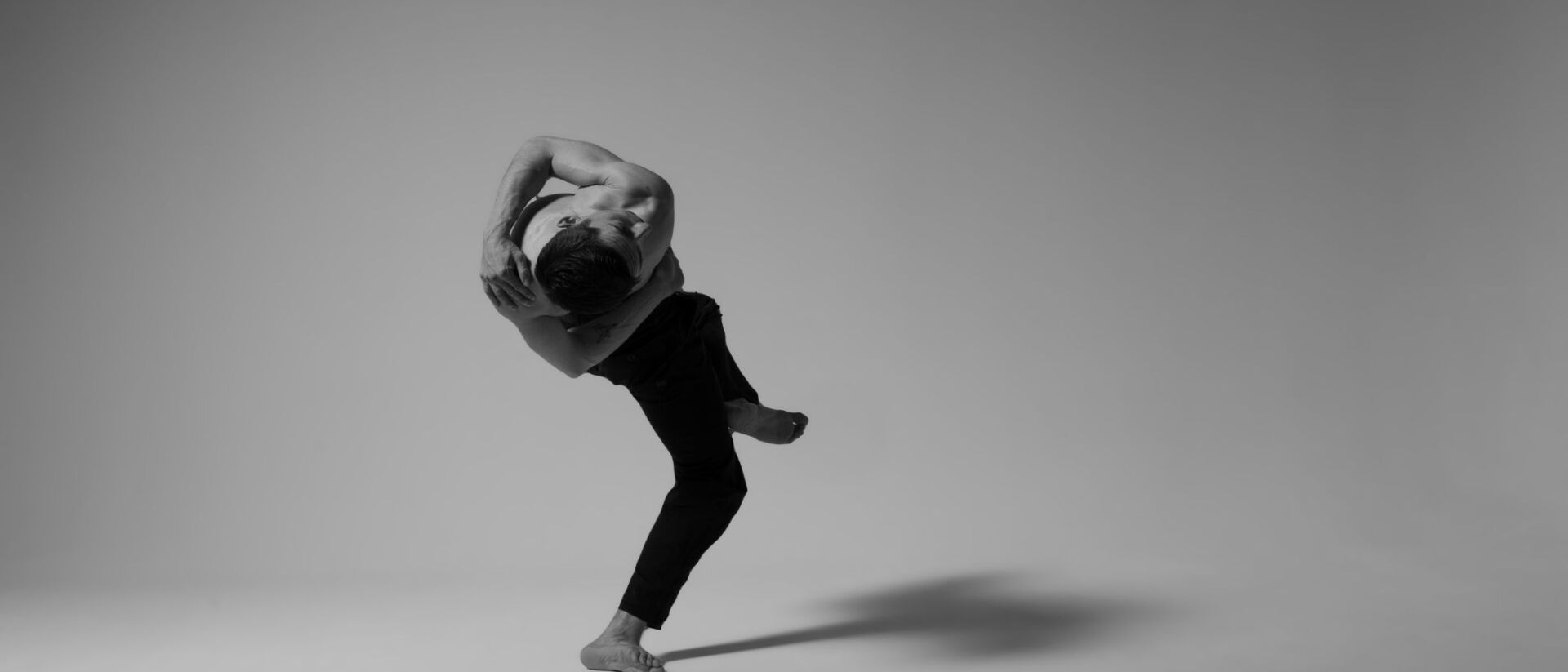 De afbeelding toont een man die een dynamische dansbeweging uitvoert, gekleed in een zwart T-shirt en broek, in een minimalistische zwart-witte setting