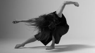 De afbeelding toont een danseres in een krachtige beweging, haar hoofd gebogen en haar haar wervelend in de lucht. Ze draagt een zwart shirt en een lange rok, dansend op blote voeten tegen een minimalistische achtergrond.