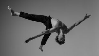 Danser in krachtige beweging met gestrekte handen en voeten, zwart-wit achtergrond.