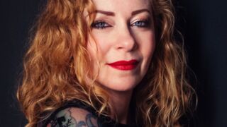 Close-up van Anneke van Giersbergen met een zachte glimlach, lang krullend haar en opvallende rode lippen.