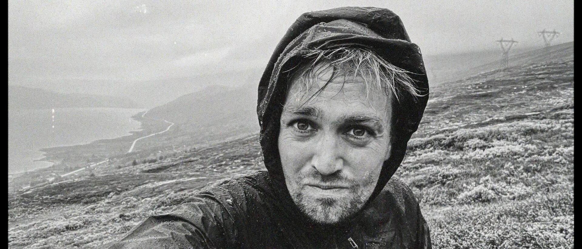 Zwart-wit selfie van Tim Knol in regenjas met capuchon, gemaakt tijdens een wandeling door een ruig, bergachtig landschap aan een kustlijn.