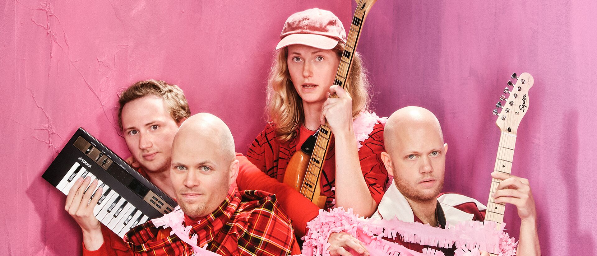 Vier personen poseren voor een roze achtergrond met muziekinstrumenten, waaronder een keyboard en twee gitaren. Ze dragen rode kleding en zijn omwikkeld met roze slingers. De sfeer is speels en kleurrijk.