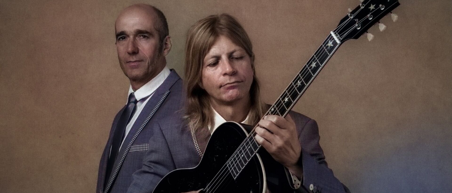 Twee personen waarvan een naar de gitaar kijkt en de ander de camera inkijkt.