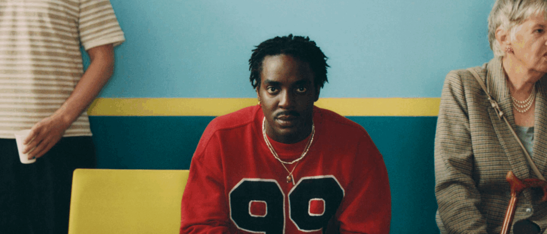 Een man met een rode trui met het cijfer 99 zit op een gele stoel in een wachtkamer met blauw-gele muren. Naast hem zit een oudere vrouw met een wandelstok, en links staat een persoon met een gestreept T-shirt en een bekertje.