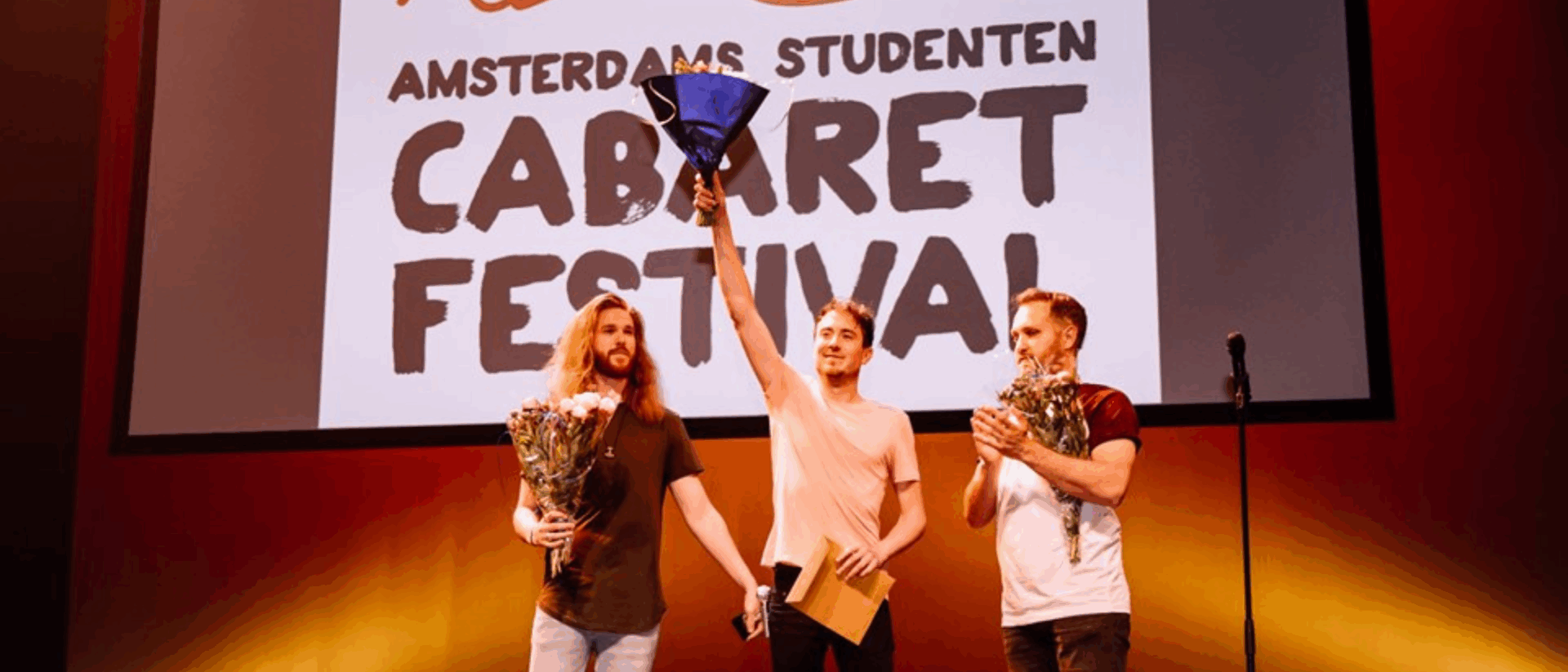 Drie mannen op een podium tijdens het Amsterdams Studenten Cabaret Festival. De man in het midden steekt juichend een boeket bloemen in de lucht; de andere twee houden ook bloemen vast. Op de achtergrond is het logo van het festival te zien.