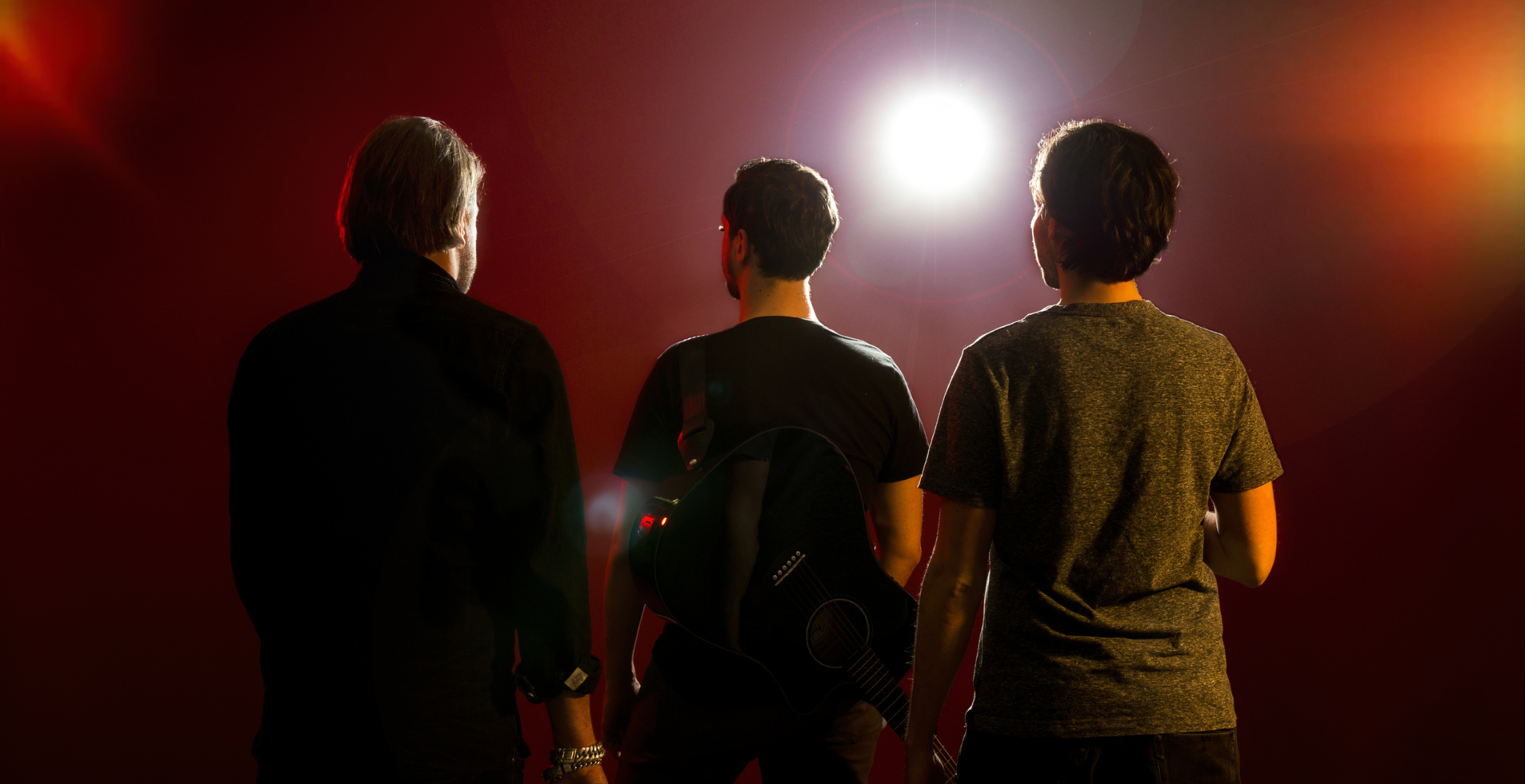 Drie jonge mannen staan met hun rug naar de camera in het schijnsel van een felle podiumspot, klaar om op te treden. Eén van hen draagt een gitaar op zijn rug. De achtergrond is donkerrood met lichte lensflares.