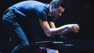 Tom Dumoulin demonstreert op het podium een tijdritpositie op een racefiets, voorovergebogen met zijn handen op het stuur, tijdens een theateravond over wielrennen.