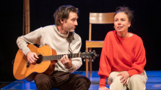 Een gitarist en actrice zitten naast elkaar op de rand van het podium.