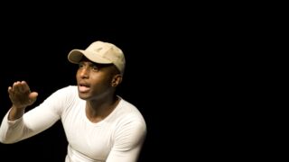 Man met wit tshirt en pet die met open mond kijkt achter hem zwarte achtergrond