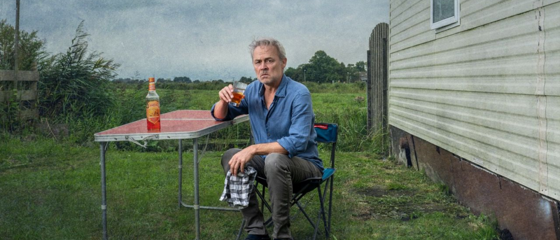 Marcel Hensema zit met glas drank aan campingtafel naast stacaravan in theatervoorstelling Eldorado