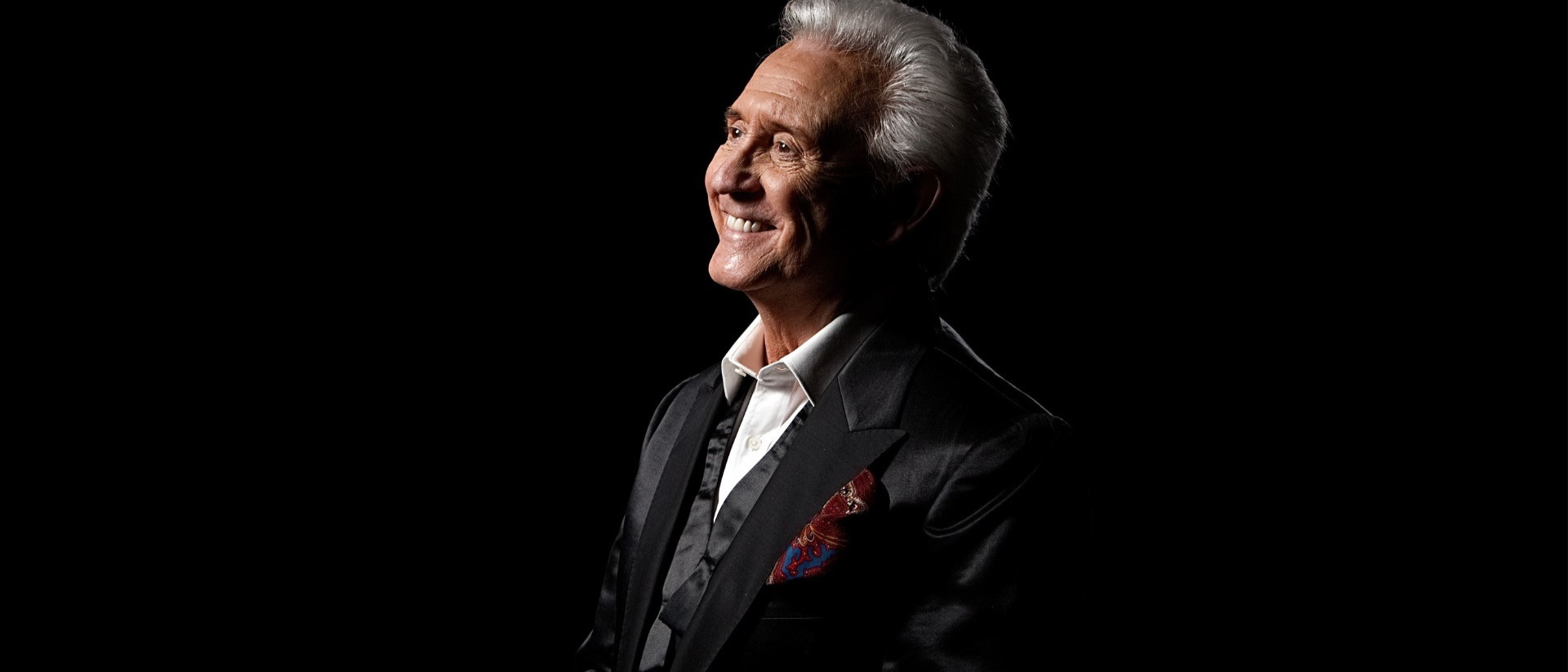 Tony Christie in zwart pak met rode pochet, lachend in spotlicht, portretfoto voor concert in Hoorn