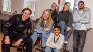 Zes jongeren die in een lokaal zitten en staan en lachend naar de camera kijken