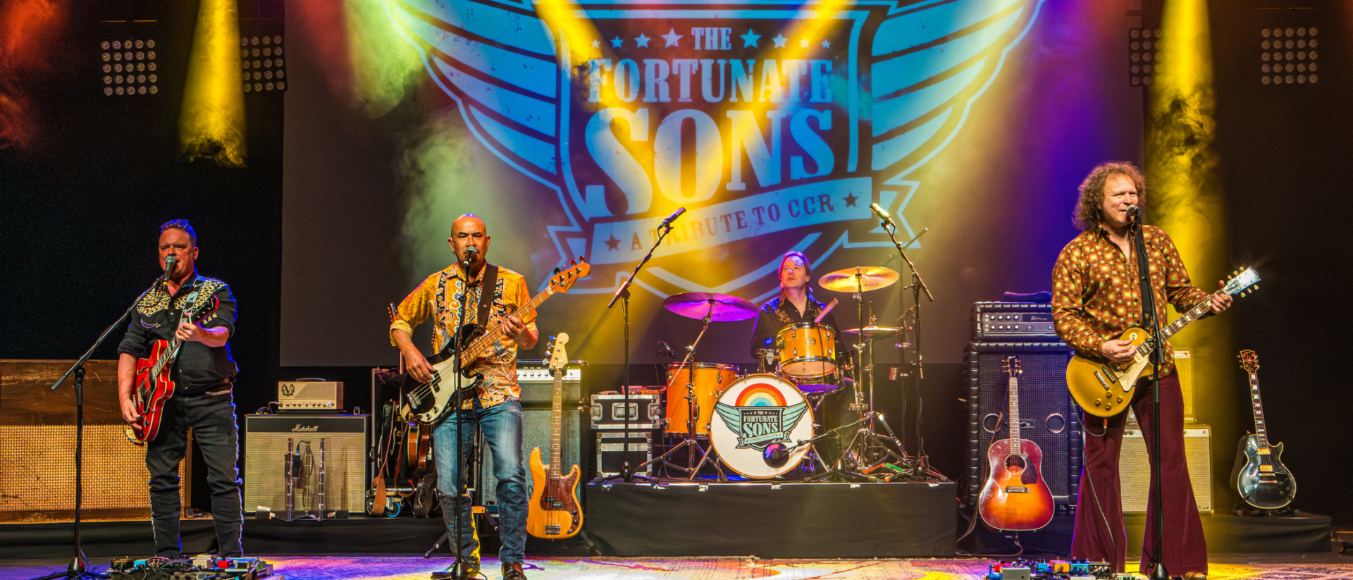 The Fortunate Sons speelt Creedence Gold op podium met gitaristen, bassist en drummer onder gekleurde theaterlichten