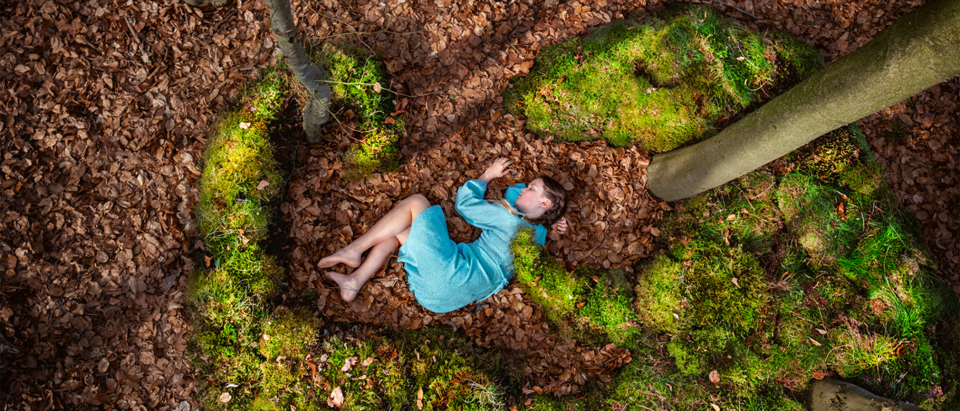 Kind in blauw jurkje liggend tussen bladeren en mos in bos, gezien van boven, Grondtonen Het Houten Huis Hoorn
